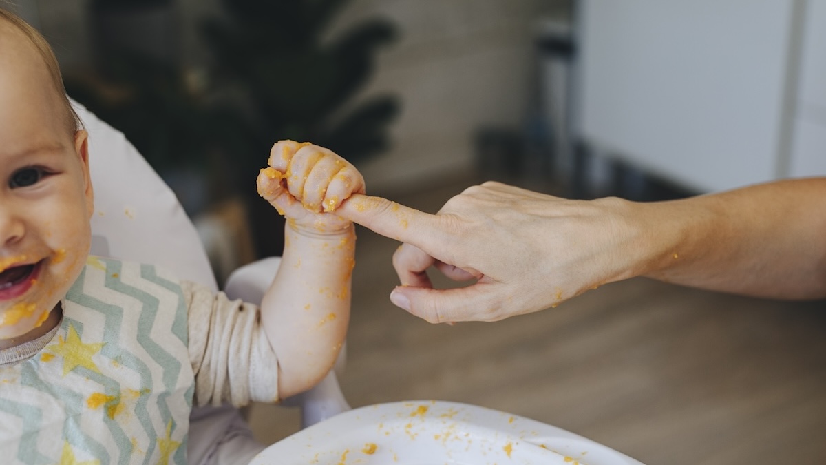 Baby mit Brei im Gesicht und an den Händen hält Finger eines Elternteils fest